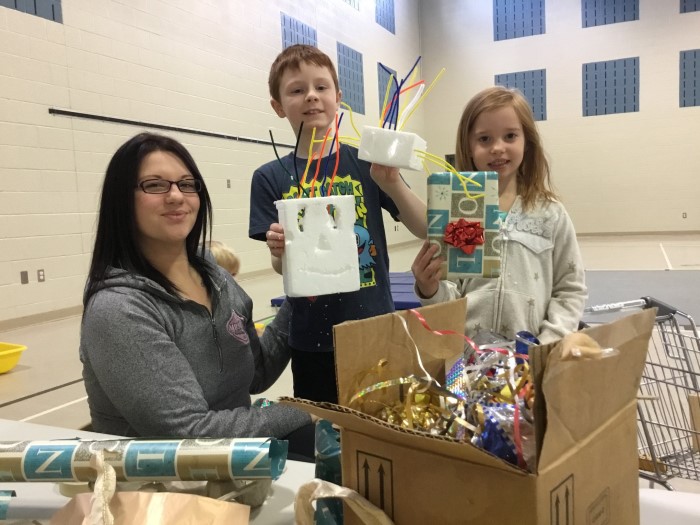 Two kids and a teacher are holding crafts in the gym of EarlyON Years Centre