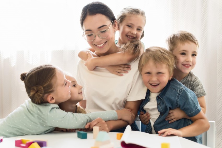 Five kids - two boys and three girls are hugging a young teacher wearing glasses